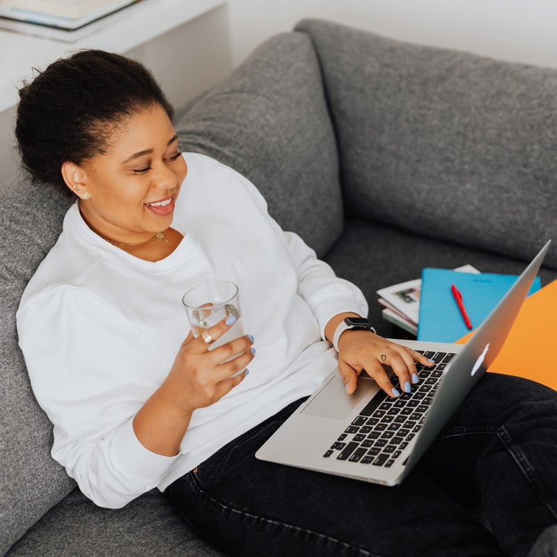 Smiling woman on a gray sofa holding a glass of water and typing on a laptop