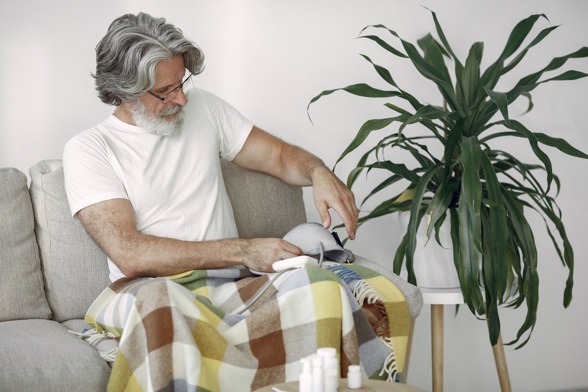 Gray-haired man adjusting a blood pressure cuff on his arm on a sofa