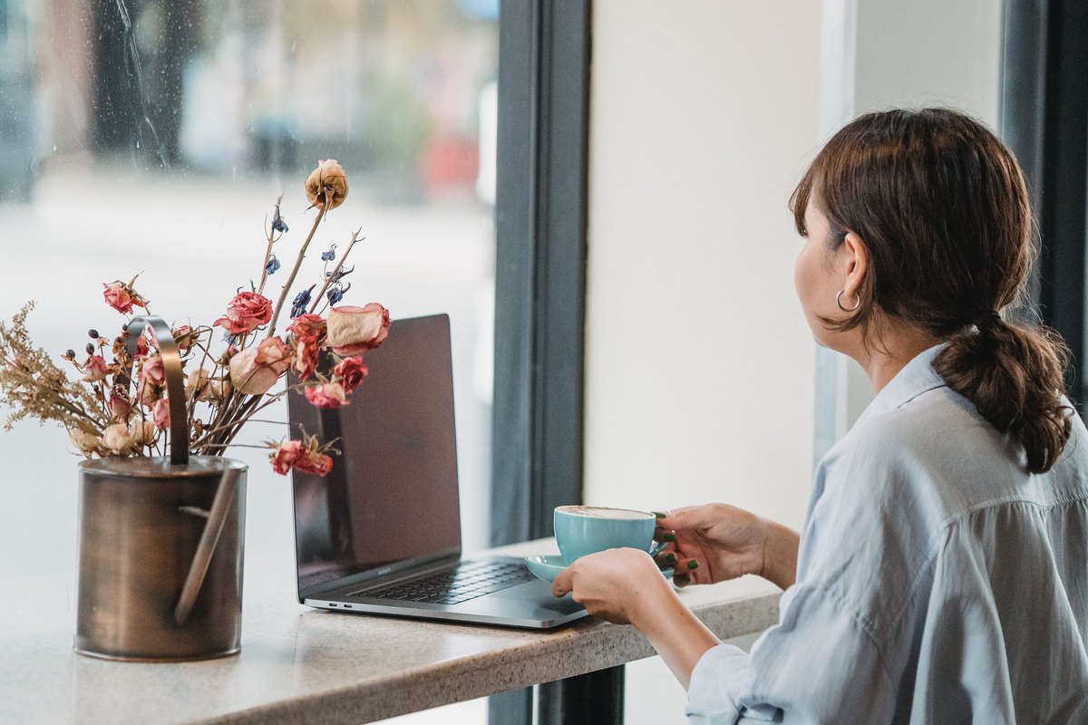 Woman holding a turquoise cup beside a laptop at a cafe window