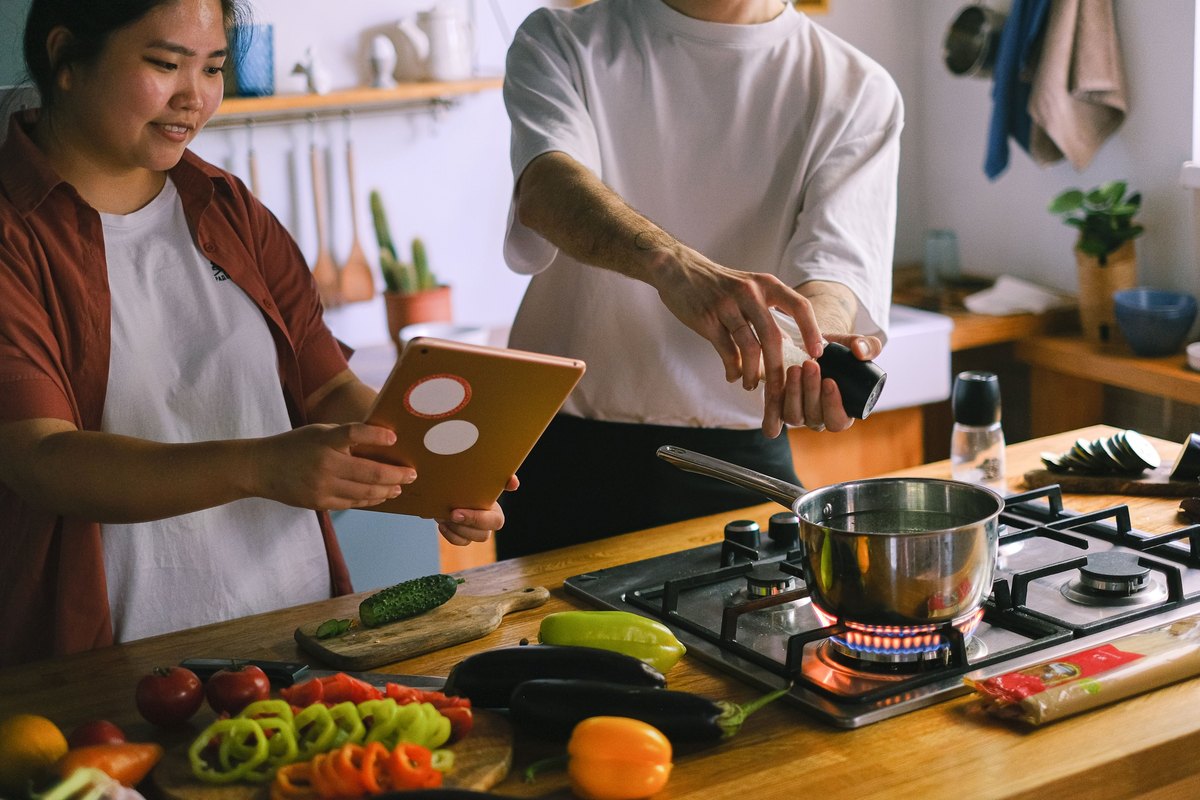 Couple cooking together, one holding a tablet recipe and one grinding pepper over a pot