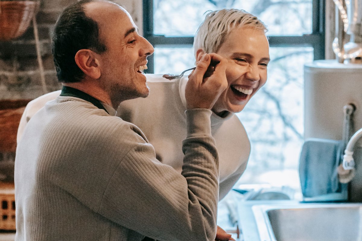 Couple laughing while sharing a taste from the pan at the stove