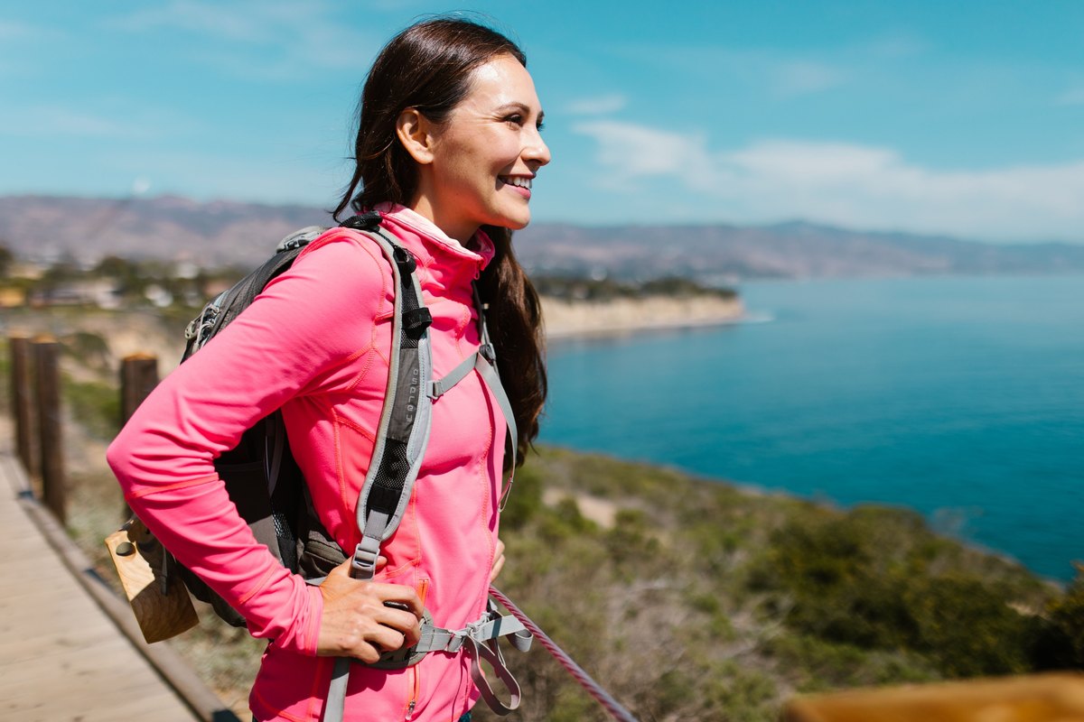 Smiling woman in a pink jacket with a daypack on a coastal boardwalk