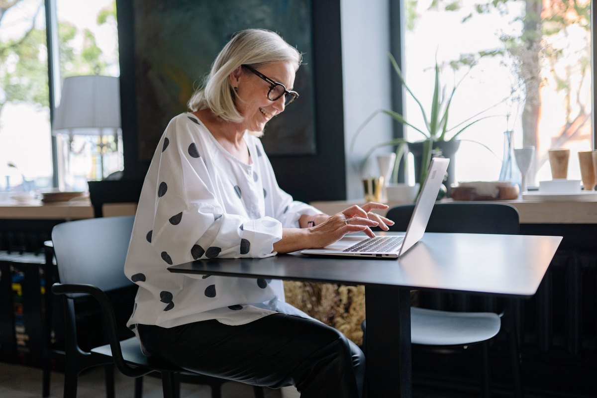 Smiling silver-haired woman typing on a laptop at a cafe table