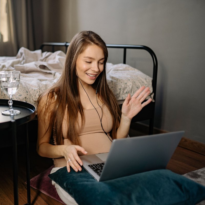 Smiling woman with earphones waving at a laptop while sitting by her bed
