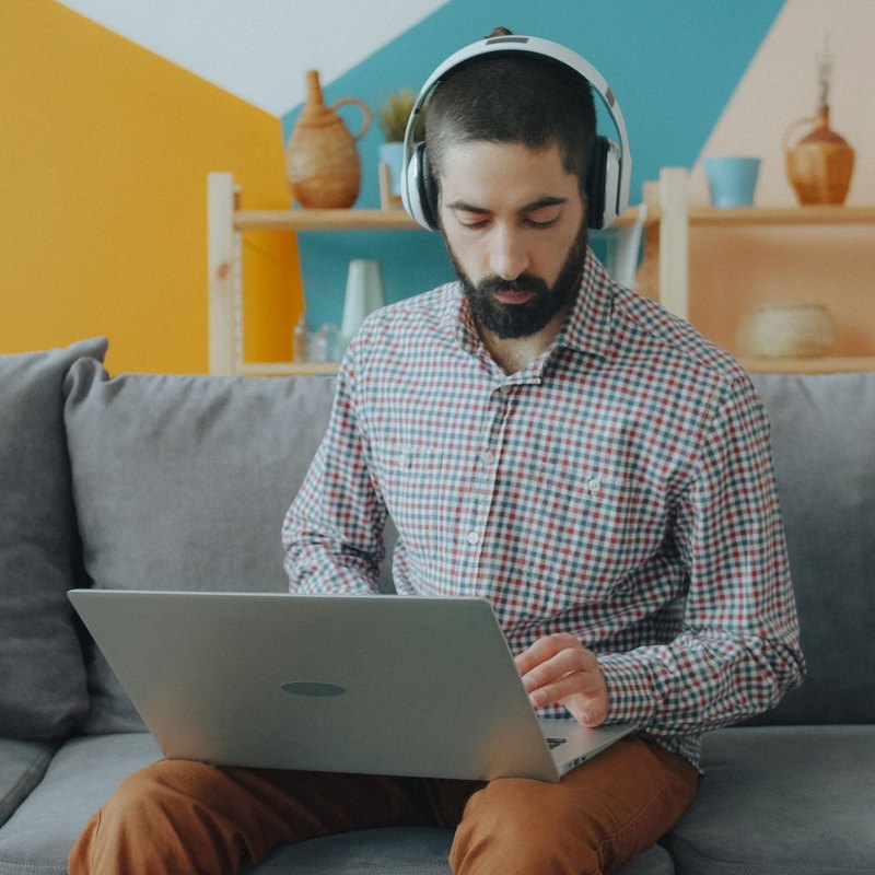 Bearded man in headphones typing on a laptop on a gray sofa