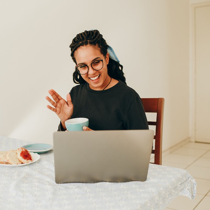 Smiling woman in round glasses waving at a laptop with a cup in her hand