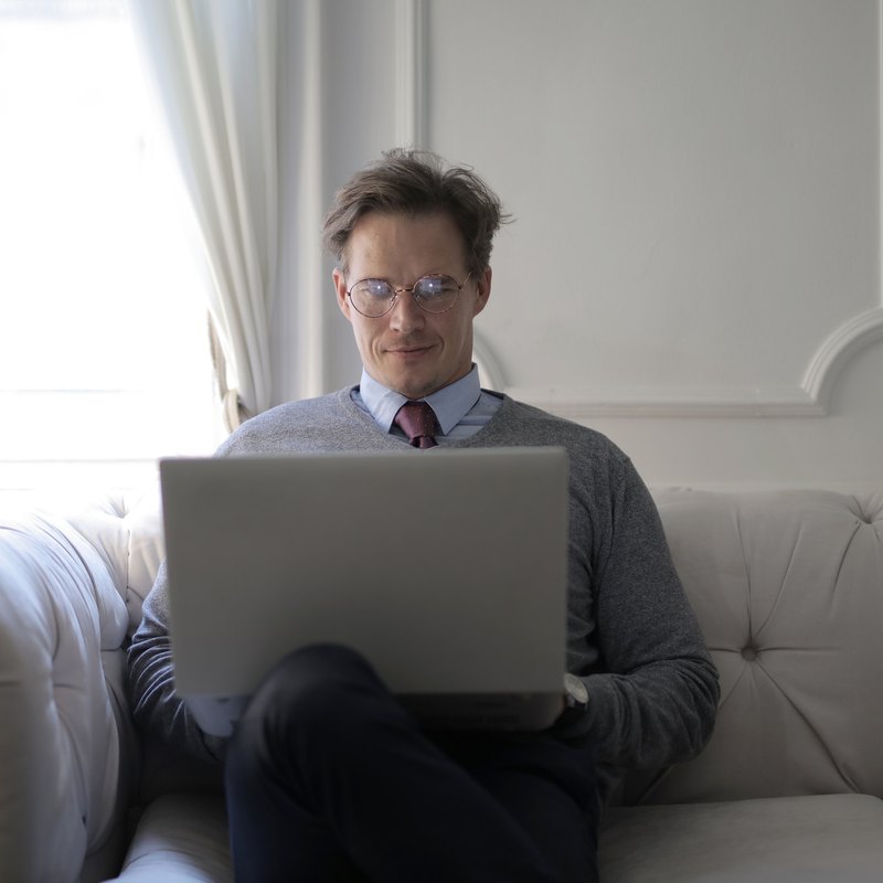 Man in round glasses smiling at a laptop on a cream sofa