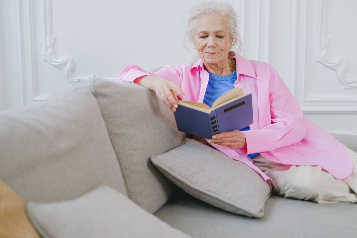 Older woman in a pink shirt reading a book on a sofa