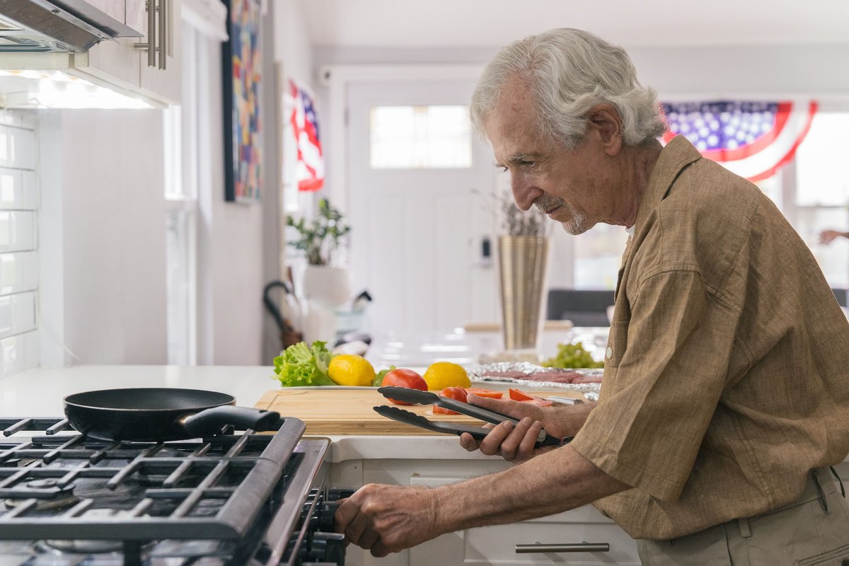 White-haired man lifting sliced tomatoes with tongs beside a stove