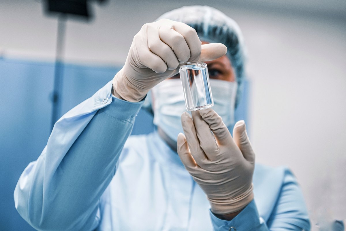 Laboratory worker in a gown and mask inspecting a small glass vial