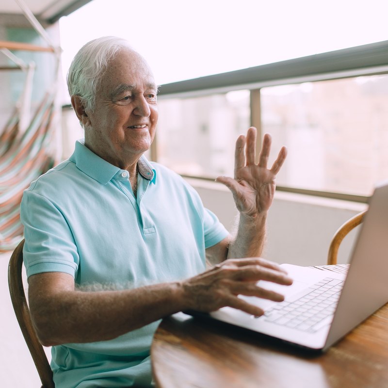 White-haired man in a blue polo shirt waving at a laptop on a balcony