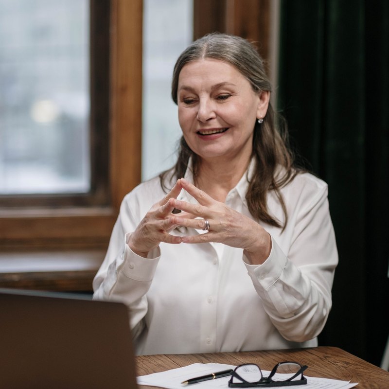 Woman with gray-brown hair smiling and talking toward a laptop at a wooden table