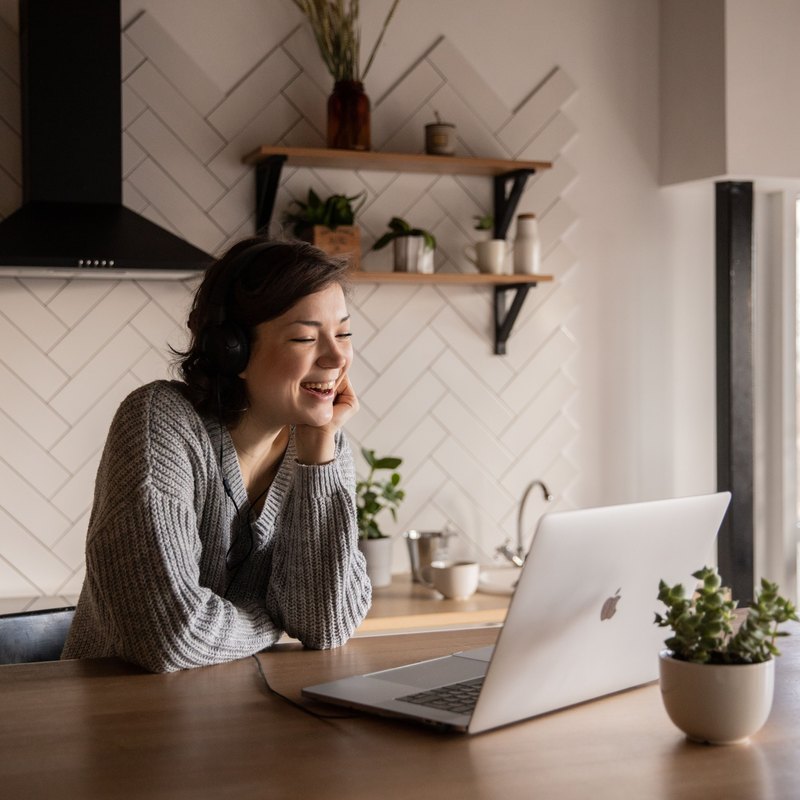 Woman with headphones smiling at a laptop on a kitchen table