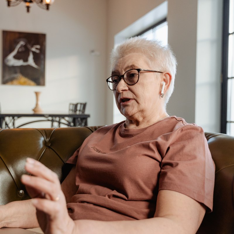 Older woman with short white hair and glasses talking toward a laptop from a leather sofa