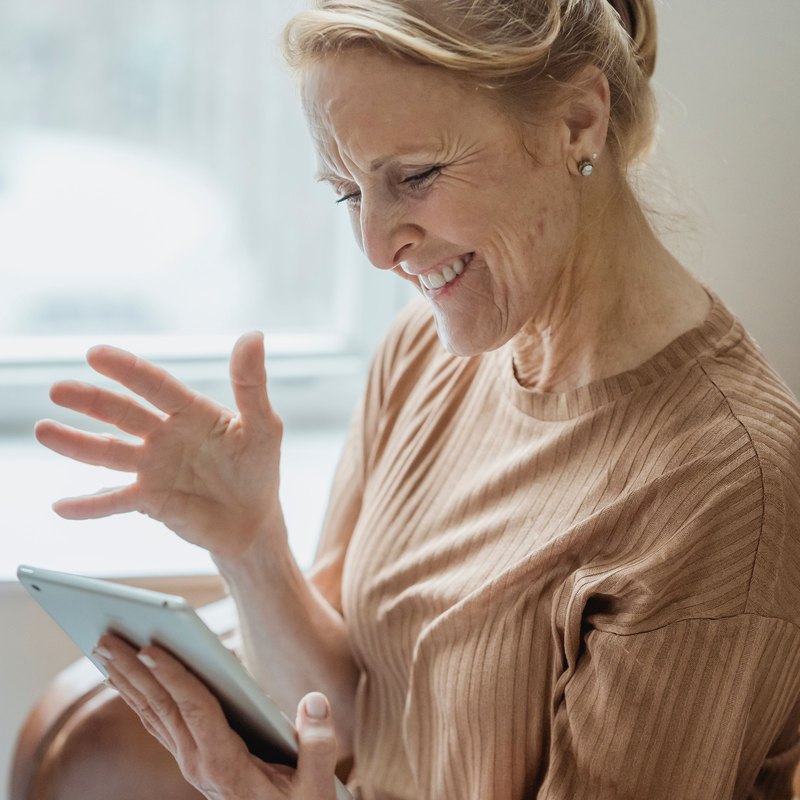 Smiling woman waving at a tablet while sitting in an armchair