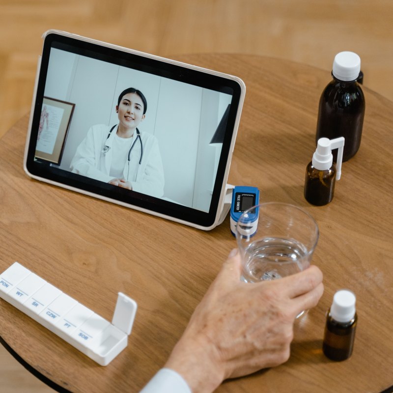 Tablet on a side table showing a woman in a white coat on a video call, beside a glass of water and medicine bottles