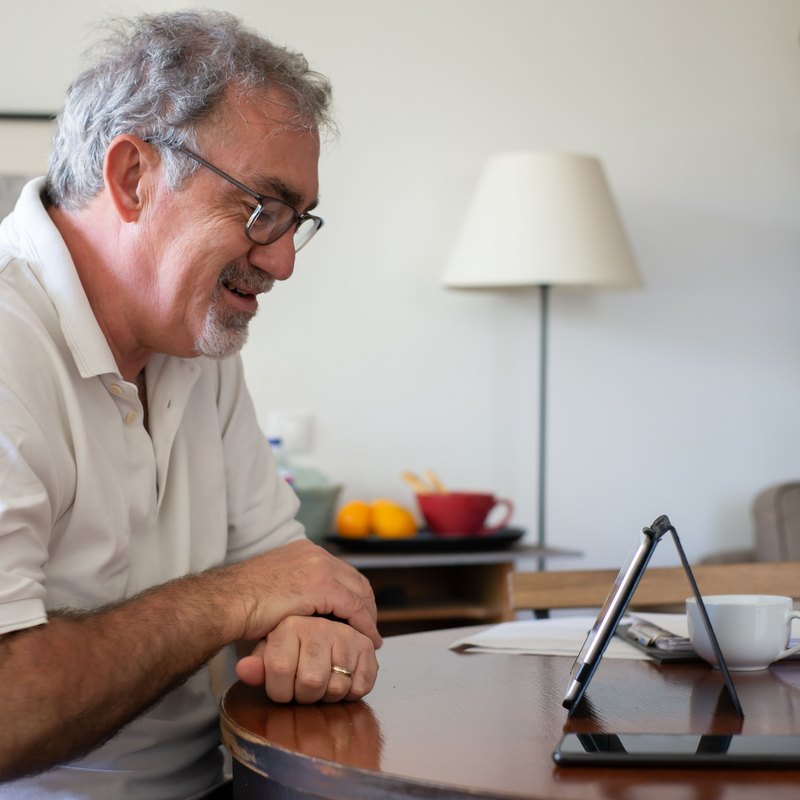 Gray-haired man with glasses smiling at a tablet on a wooden table