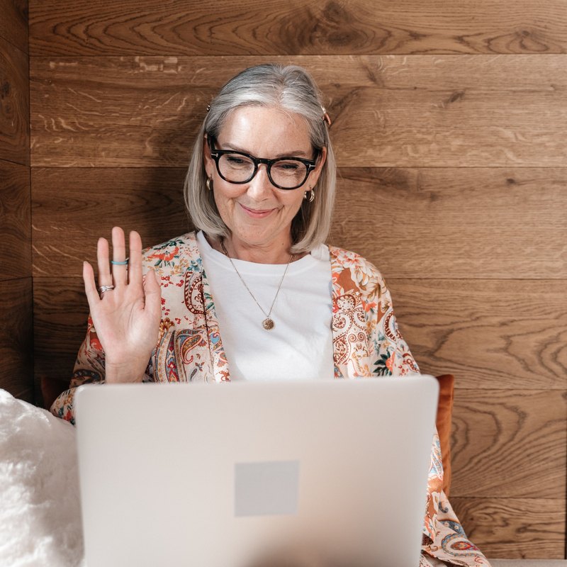 Gray-haired woman in glasses smiling and waving at a laptop
