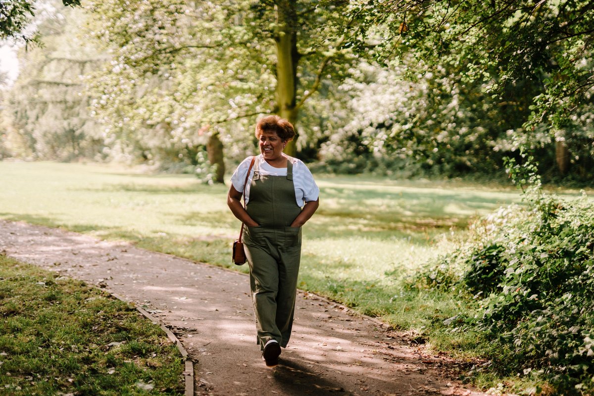 Woman in olive overalls walking along a tree-lined park path