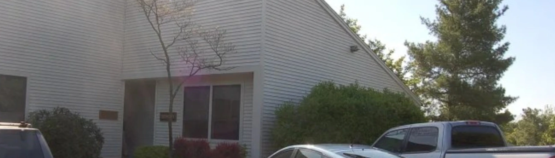 Single-story gray office building with large windows, trees and parked cars out front